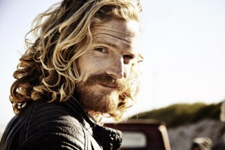 man with long hair standing in the wind close to the ocean, looking into camera