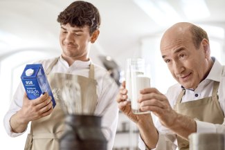 Two men in Weihenstephan aprons in a kitchen – one holds a Weihenstephan Haltbare Milch carton, the other inspects a glass of milk with a smile.