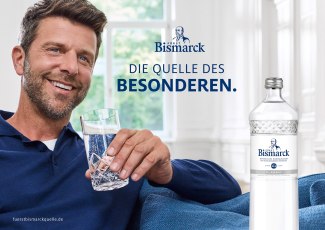 A relaxed smiling man holds a glass of Fürst Bismarck Classic mineral water on a sofa next to a Fürst Bismarck bottle with the tagline "Die Quelle des Besonderen."