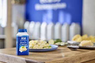 A Weihenstephan Koch-Creme bottle stands on a wooden kitchen counter next to a plate of creamy pasta with courgette, the Weihenstephan logo visible in the background.