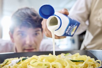 Weihenstephan Koch-Creme being poured from its blue bottle onto a pan of tagliatelle with courgette while a young man watches intently in the background.