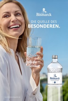 A laughing blonde woman holds a sparkling glass of Fürst Bismarck mineral water outdoors under a clear blue sky next to a Fürst Bismarck Classic bottle with the tagline "Die Quelle des Besonderen."
