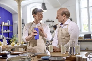Two men in Weihenstephan aprons stand at a kitchen island opening a Weihenstephan milk carton, surrounded by glasses of milk and festive Christmas decorations