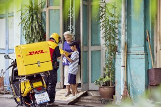 A DHL motorcycle courier hands over a parcel to an elderly woman and a young boy at a colorful doorstep in Bangkok