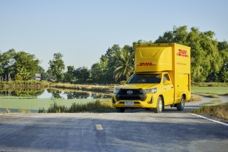 A yellow DHL delivery truck drives along a quiet rural road beside a peaceful pond in the Thai countryside