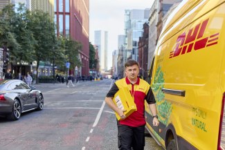 A DHL courier carries parcels along a busy Manchester city street next to a yellow DHL electric delivery van