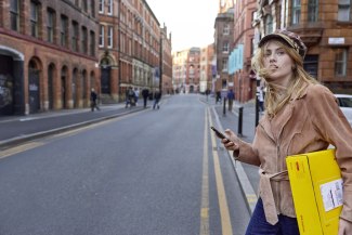 A young woman holding a DHL Express parcel and her smartphone looks over her shoulder on a Manchester red brick street