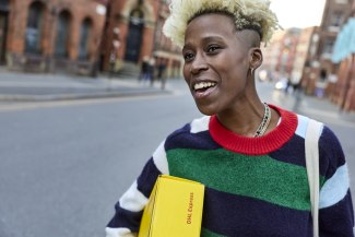 A smiling young woman carries a yellow DHL Express parcel on a Manchester street lined with red brick Victorian buildings.