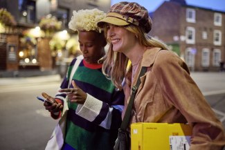 Two young women check a smartphone together on an illuminated Manchester street at night, one carrying a yellow DHL parcel