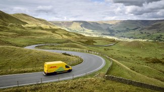A yellow DHL electric van winds along a sweeping mountain road through the rolling green hills of the Peak District.