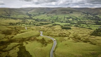 Aerial view of a yellow DHL van on a winding country road cutting through vast moorland and valley landscape.