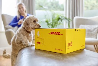 A fluffy golden dog sits next to a yellow DHL Express parcel on a sofa while a woman smiles in the background.