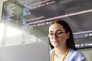 A focused female DHL employee works on a laptop in front of a large screen displaying programming code.