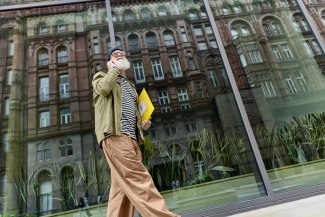 A stylish older man walks past a glass facade reflecting Manchester's Victorian architecture, carrying a yellow DHL envelope while on the phone