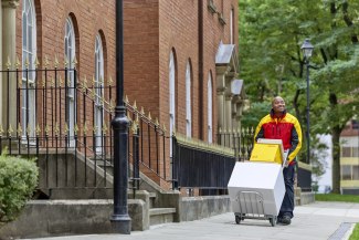 A smiling DHL courier pushes a trolley loaded with parcels along a pavement in front of a classic red brick Manchester building.