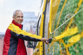 A smiling male DHL courier opens the door of his yellow electric delivery van on a Manchester street.
