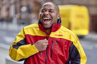 A laughing DHL courier zips up his jacket in front of a yellow DHL van on a Manchester street.
