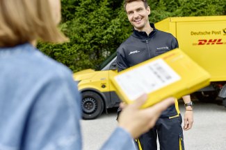 A smiling DHL courier hands a yellow parcel to a customer in front of a Deutsche Post DHL delivery van.