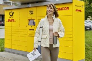 A smiling woman stands in front of a Deutsche Post DHL Packstation holding a letter and a bag after collecting her shipment