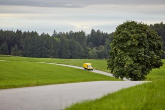 A yellow Deutsche Post delivery van drives along a winding country road through green fields and forests.