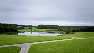 A yellow Deutsche Post van drives along a rural road past a pond and rolling green hills under a cloudy sky