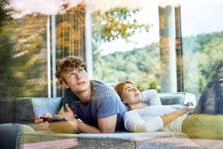 young couple laying on the sofa, controlling their smart homew with mobile phone