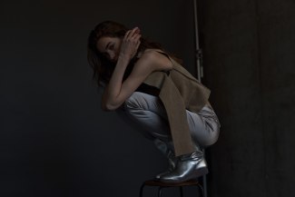 woman cowers on a stool with dark cloths in a dark environment 