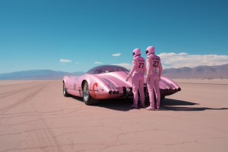 two men with pink helmet and pink overal standing beside their racingcar somewhere in a desert